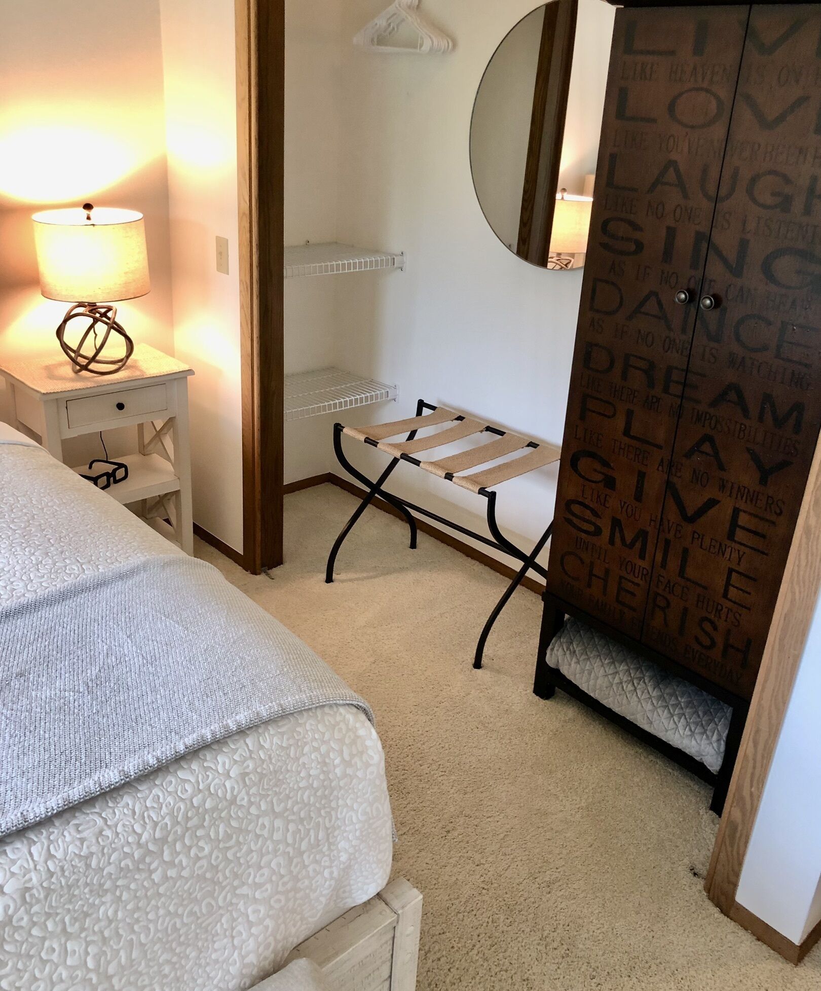 A bedroom with a bed, a bedside lamp, a small table, a chair, a mirror, and a wardrobe with motivational quotes.