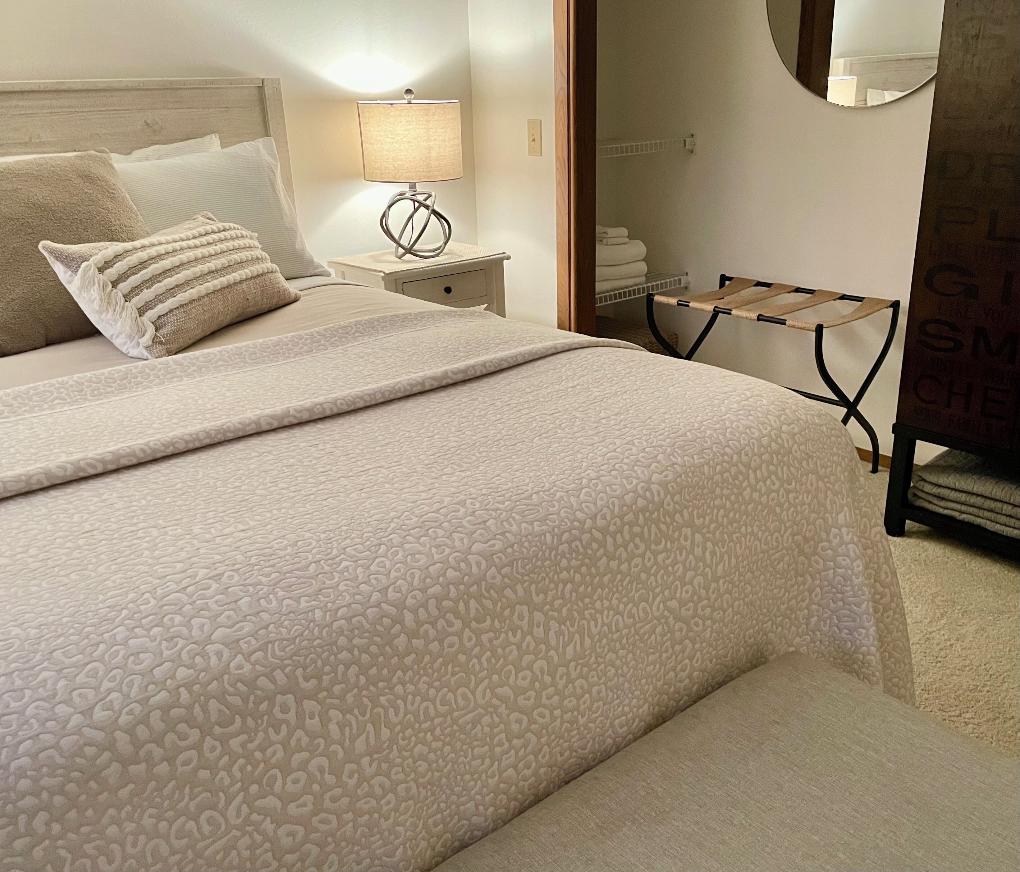 Una cama bien hecha con una colcha estampada, una lámpara de noche y una silla de madera con un perchero.