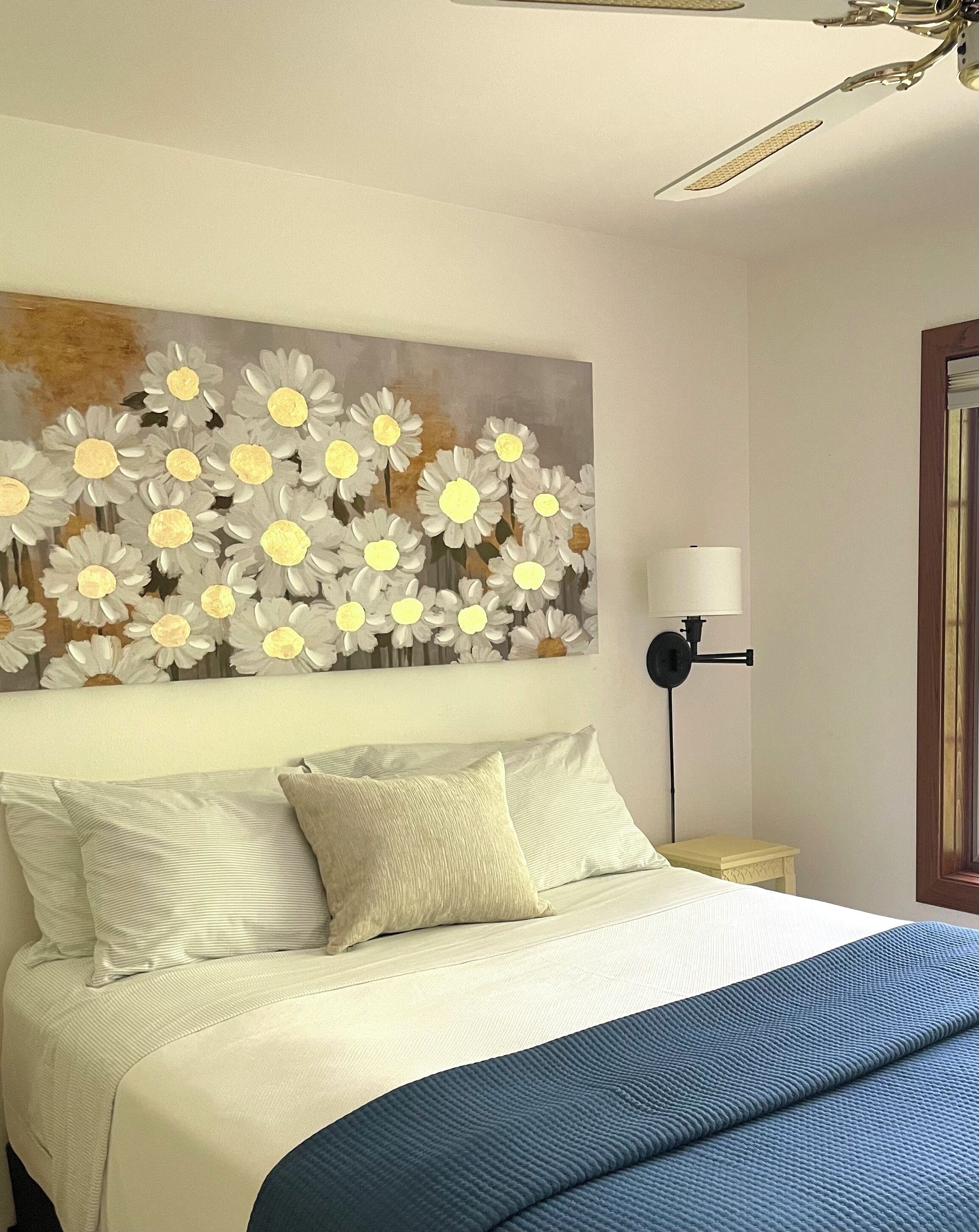 Et soveværelse med en seng, puder, et natbord, en lampe og et maleri af margueritter på væggen.