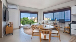 Un salon moderne avec une table ronde en bois et des chaises, un canapé et un téléviseur. De grandes fenêtres offrent une vue sur la ville et l’océan.