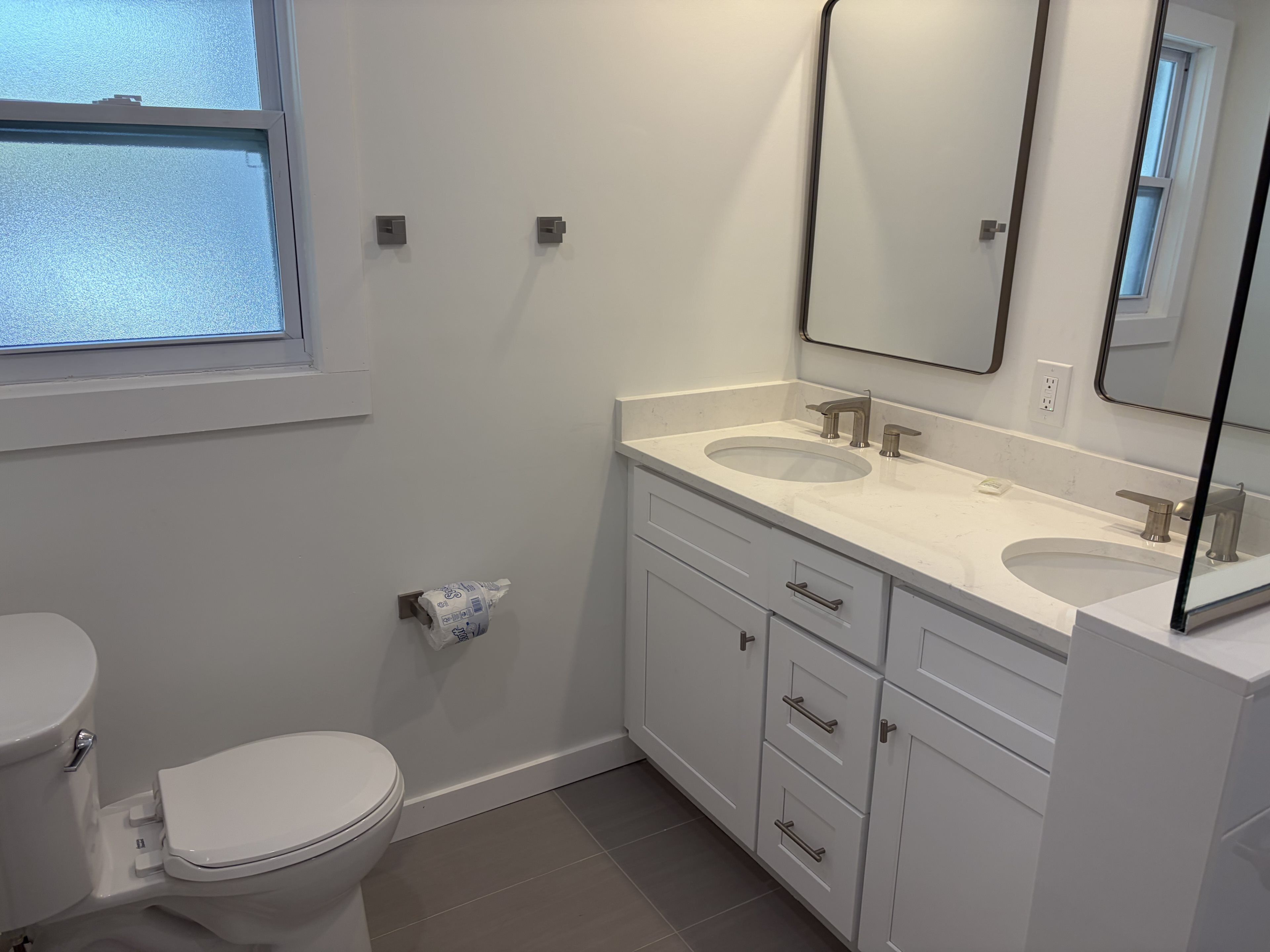A modern bathroom with a toilet, two sinks, a mirror, and a window.