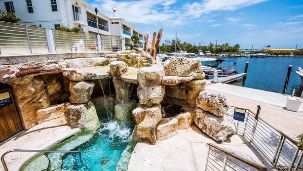 A pool with a waterfall feature, surrounded by stone structures, with boats docked in the background and a building to the left. - FULL TIME MGR - LUXURY PENTHOUSE - OFFERS KEY LARGO'S BEST PANORAMIC OCEAN VIEWS (Key Largo)