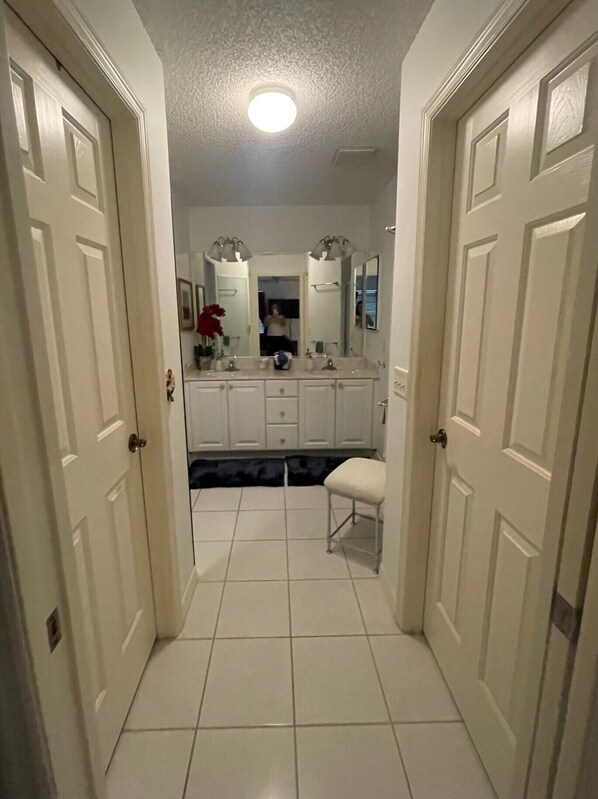 A hallway leading to a bathroom with white tiled floors, a sink, and a mirror.