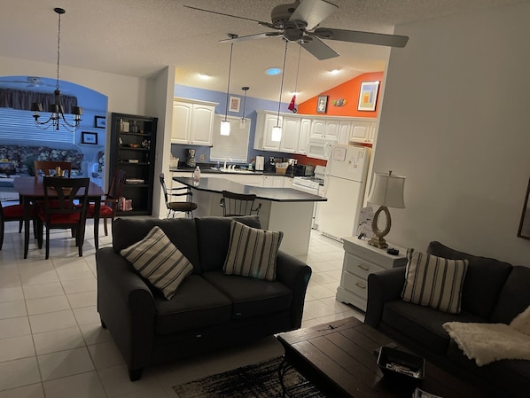 A modern living room with a dark sofa set, a coffee table, and a dining area with a table and chairs. A kitchen with white cabinets and a small island is visible in the background.
