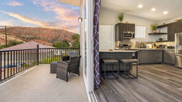 A modern kitchen with a bar area, a balcony with outdoor seating, and a scenic mountain view. - The Fortress (Cedar City)