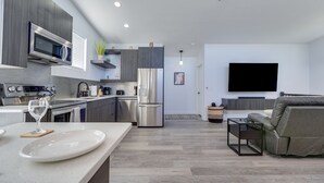 A modern kitchen with a microwave, stove, and refrigerator. There is a dining area with a white table and a glass of water. A gray sofa is in the living room with a TV mounted on the wall. - The Fortress (Cedar City)