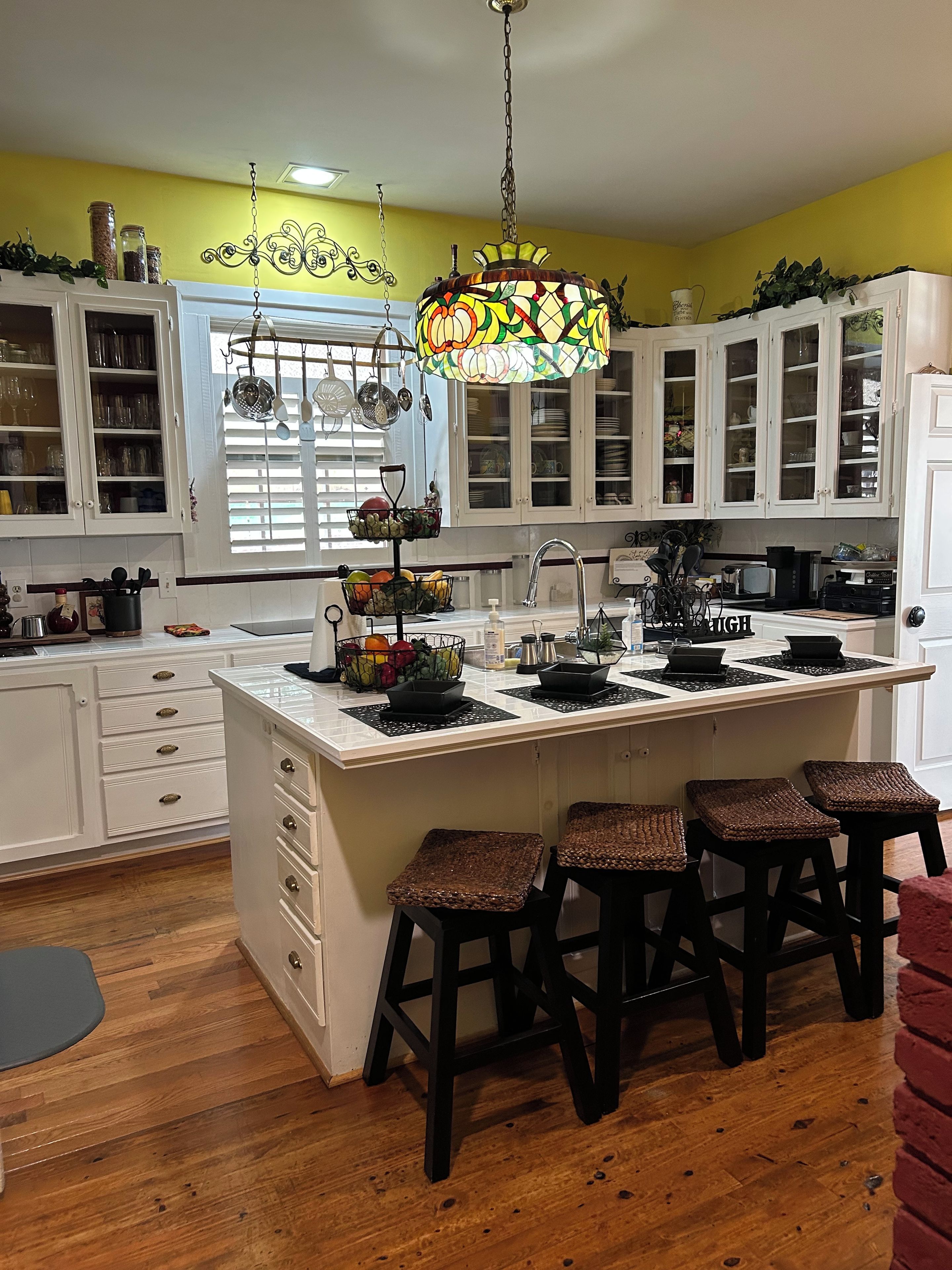 A kitchen with white cabinets, a central island with stools, a hanging chandelier, and a window with blinds.