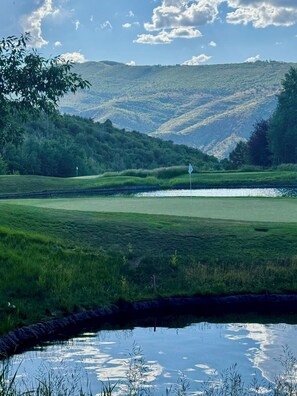 A golf course with a pond in the foreground, surrounded by lush greenery and hills in the background.