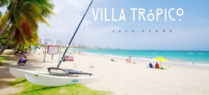 Une scène de plage avec un bateau, des palmiers et un ciel dégagé. Le texte indique : « VILLA TRÓPICO ISLA VERDE ».