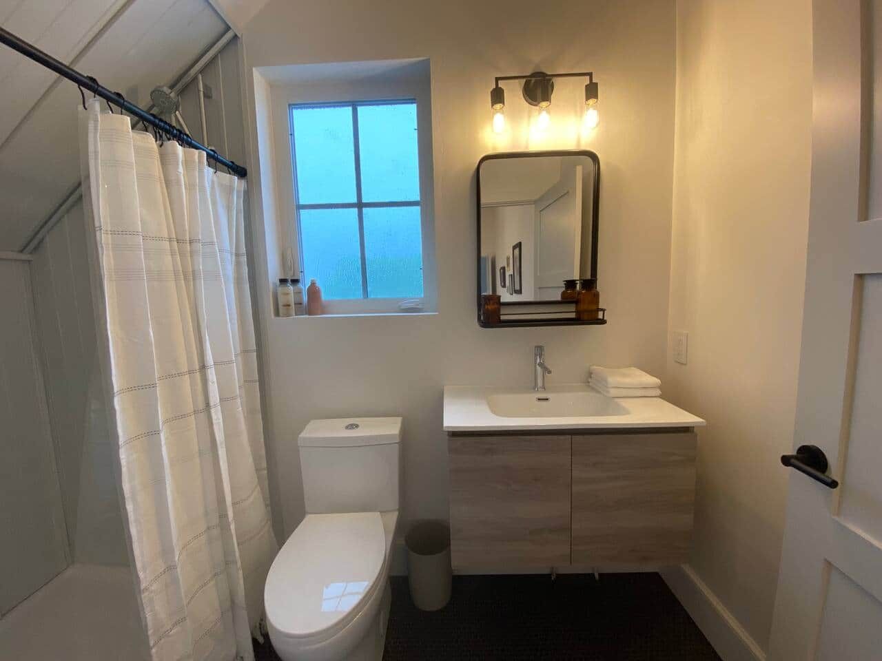 Un baño con cortina de ducha, inodoro, lavabo con espejo y ventana.