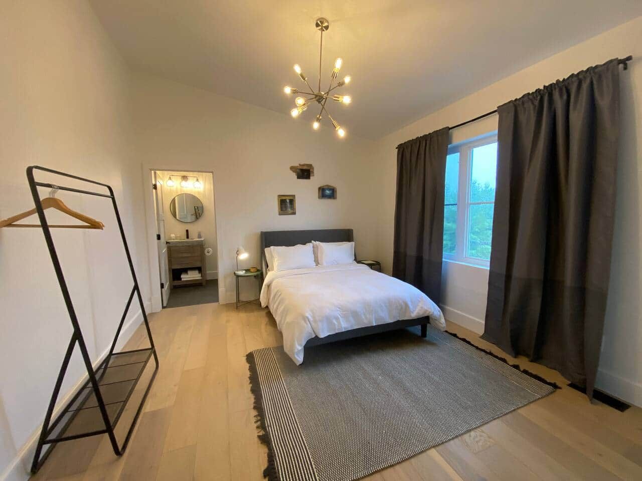 Un dormitorio con cama, mesitas de noche, lámpara colgante, ventana con cortinas y un espejo.