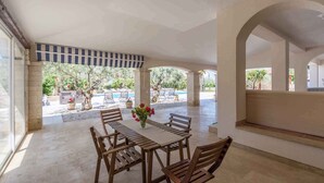 Un patio al aire libre con una mesa y sillas, rodeado de columnas y vistas a una piscina y follaje.