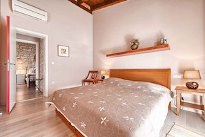 Une chambre à coucher comprenant un lit, des tables de chevet, une chaise, une étagère en bois ornée d’objets décoratifs et une porte donnant sur une autre pièce.