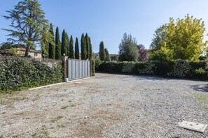 Une allée de gravier mène à une entrée fermée par un portail, le tout étant entouré de différents arbres et arbustes.