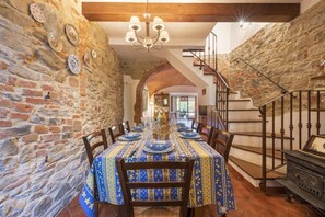 Un espace repas agrémenté d’un mur en pierre, de poutres apparentes en bois au plafond, d’un lustre et d’un escalier.