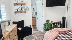 Ein modernes Schlafzimmer mit einem Bett, einem Sessel, einer Holzkommode, einem Fernseher und einer Küche im Hintergrund.