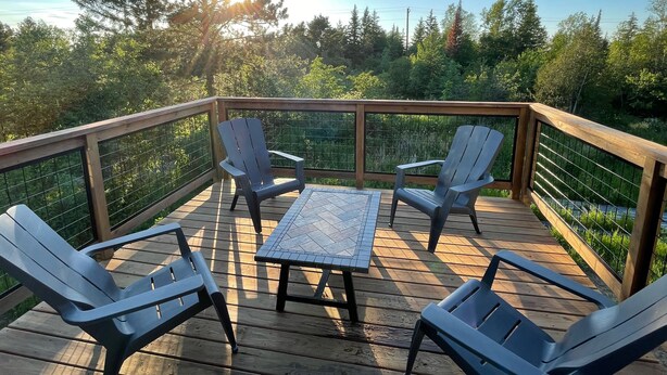A wooden deck with four Adirondack chairs and a small table, surrounded by a wooden railing and a metal fence. - Spacious 3 bedroom 2 bath lodging near trails, with lots of off-street parking! (Ely)