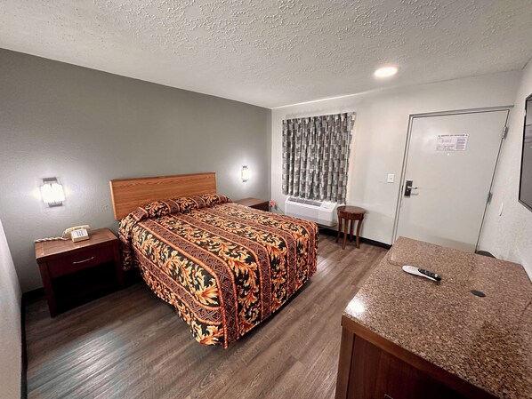 A hotel room with a bed, bedside table, nightstand, and a small desk with a chair.