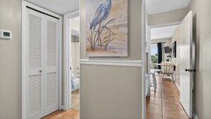 A hallway with a white door, a painting of a bird, and a view into another room with a chair and a table.