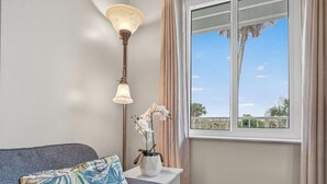 A room with a bed, a bedside table with a lamp, a window with curtains, and a view of palm trees.