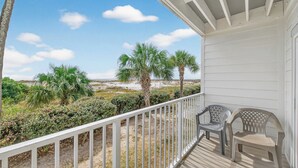 A covered porch with a white railing, two plastic chairs, and a view of the beach and palm trees.