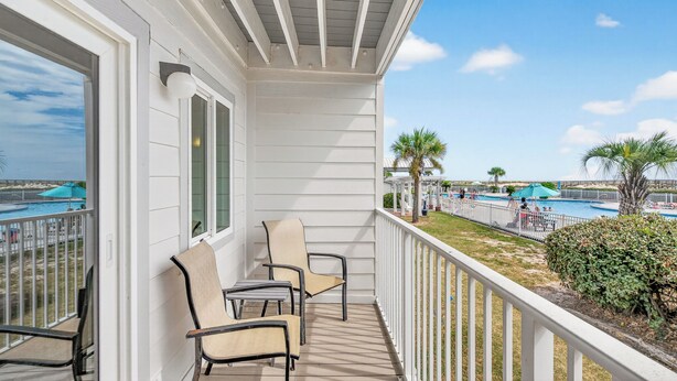 A balcony with white railings, beige chairs, and a view of a pool area with umbrellas and palm trees.