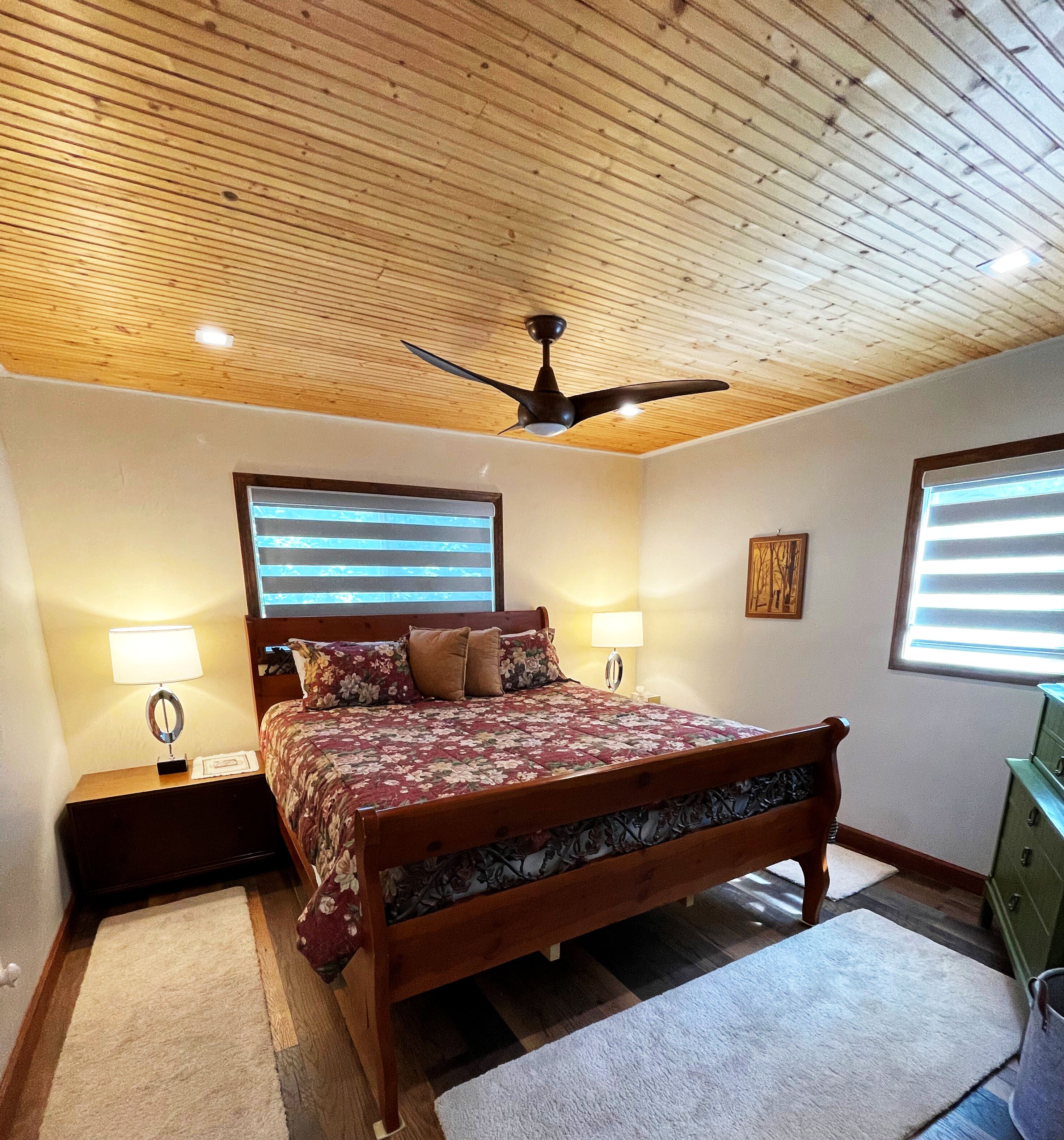 Un dormitorio con techo de madera, una cama con ropa de cama floral, dos lámparas de noche y un ventilador de techo.
