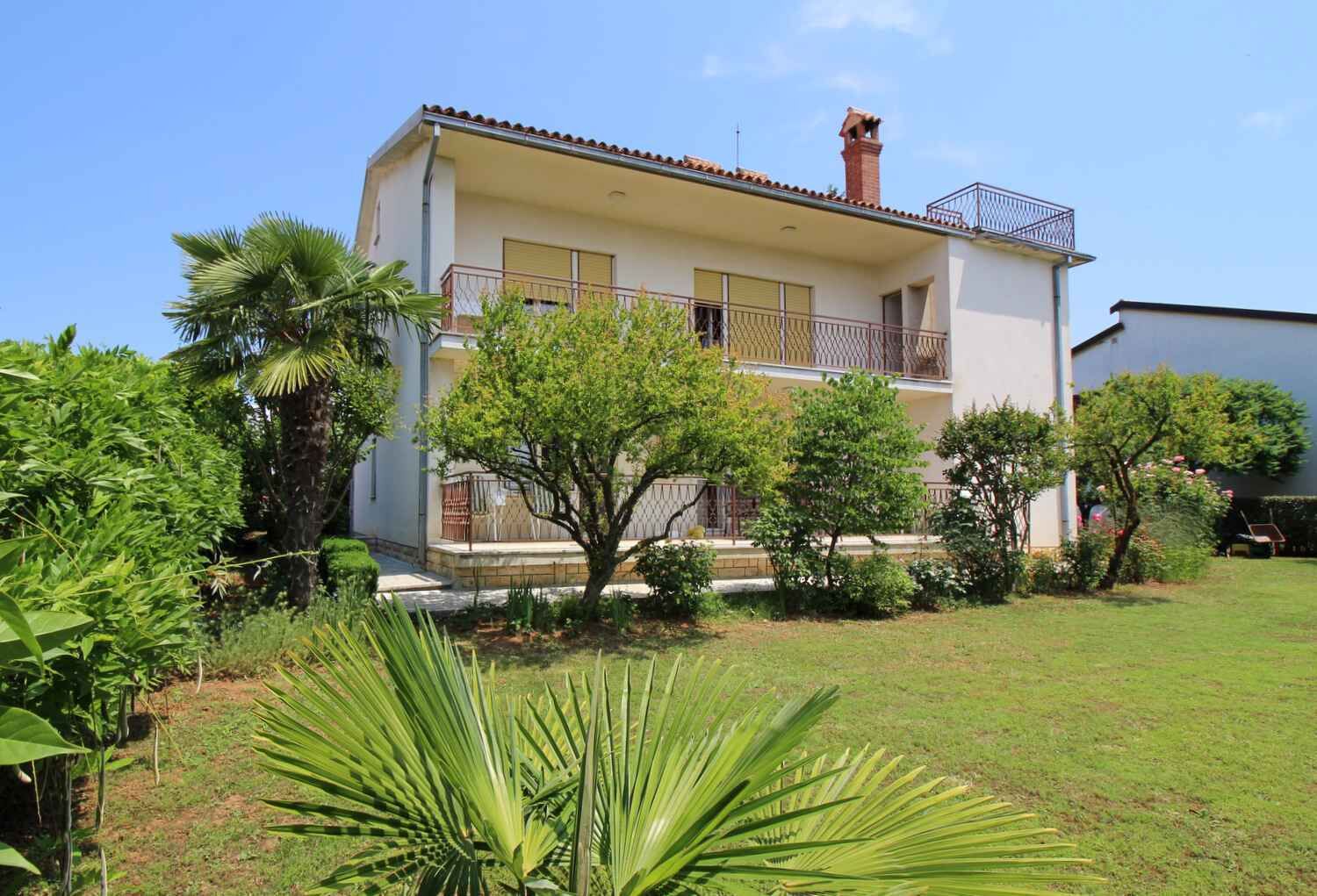 A two-story building with a balcony, surrounded by greenery and a well-maintained lawn.
