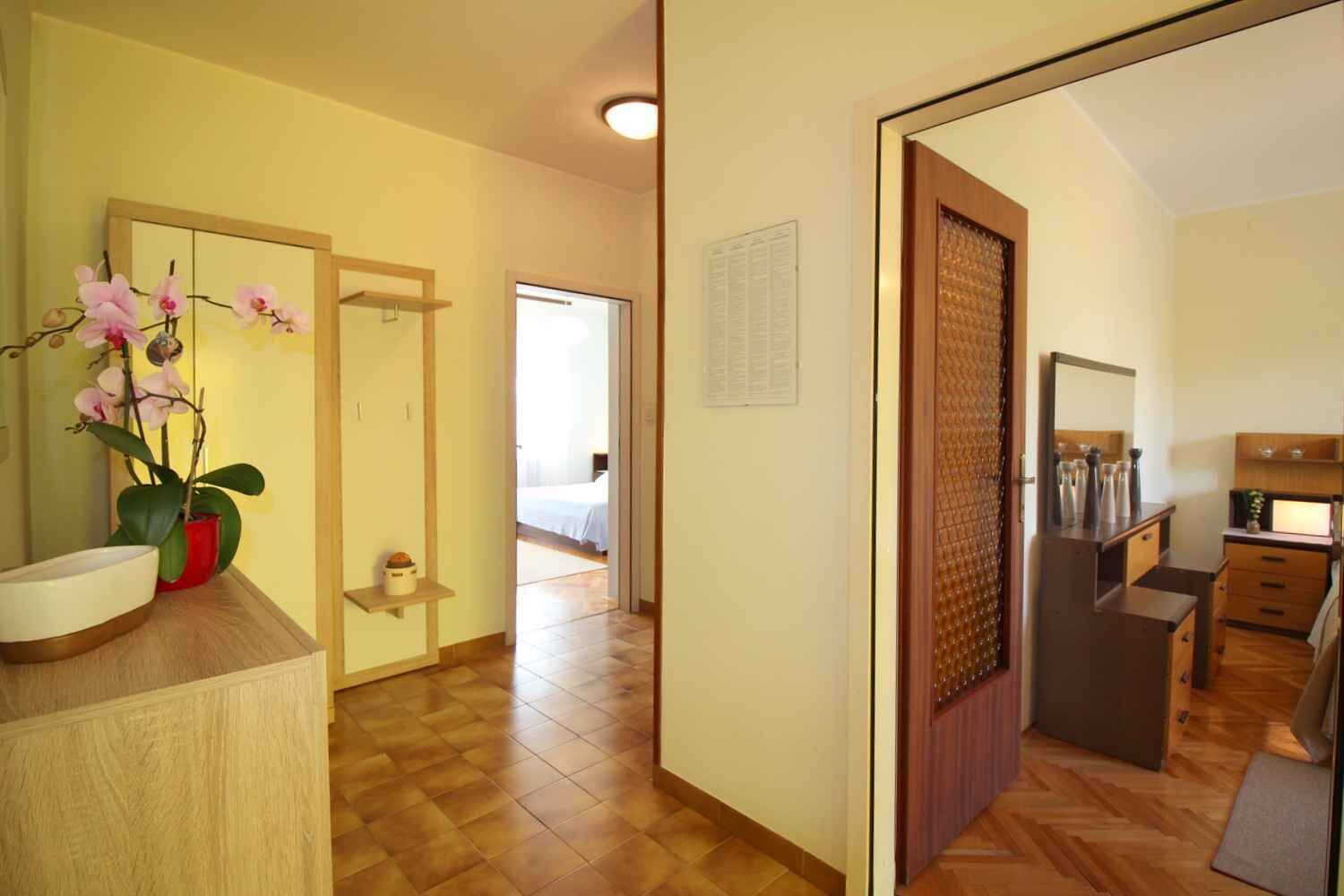 A hotel room with a wooden floor, a bed, a small table with a vase, a mirror, and a dresser.