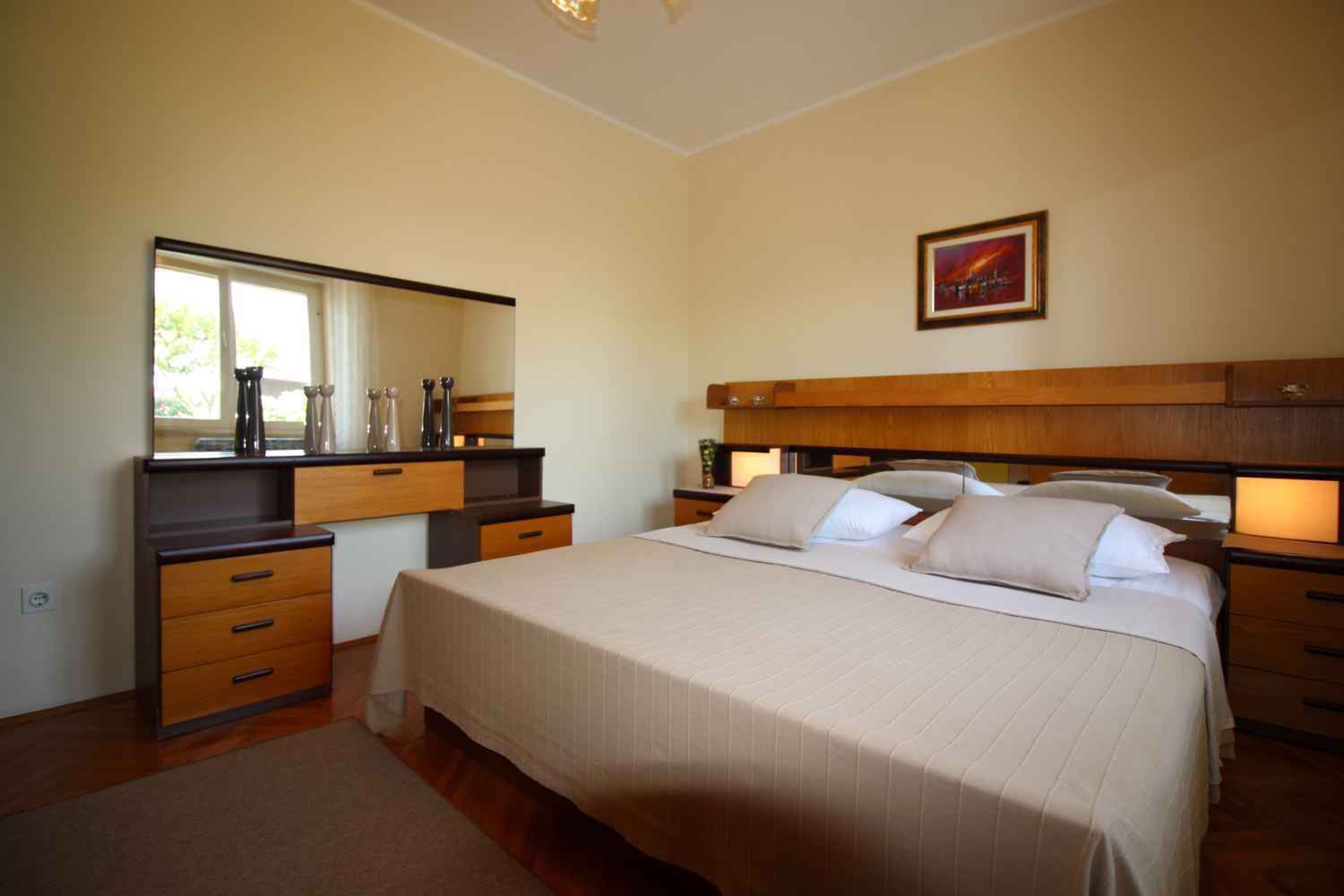 Een slaapkamer met een bed, nachtkastjes, een dressoir, een spiegel en een lijst met een afbeelding aan de muur.