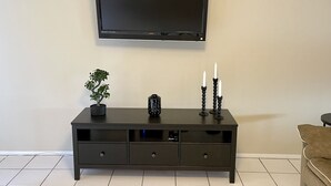 Un meuble TV en bois foncé, surmonté d’un téléviseur à écran plat, avec une petite plante en pot et deux chandeliers contenant des bougies blanches.