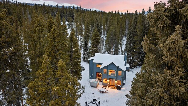 A large cabin surrounded by a dense forest, with a snow-covered roof and a visible deck area.