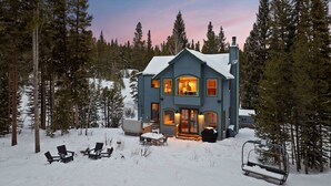 A two-story house with a snow-covered roof, surrounded by tall pine trees.