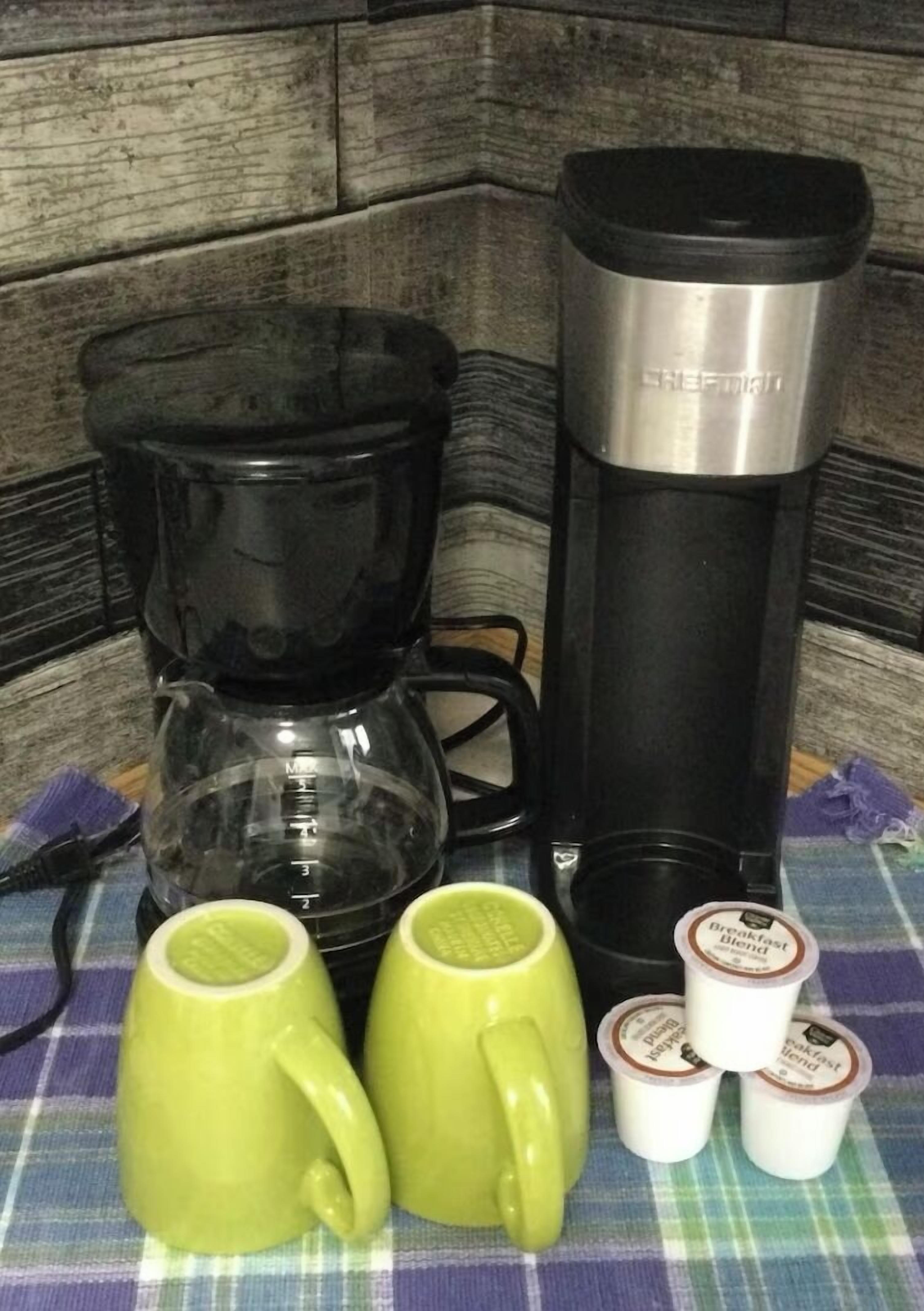A kitchen area with a coffee maker, a thermos, and a set of mugs and containers on a checkered tablecloth.