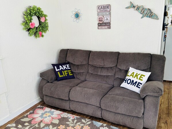 Un sofá marrón con dos almohadas, un tapete floral y adornos de pared que incluyen un lazo, un pez y letreros con motivos de cabaña.