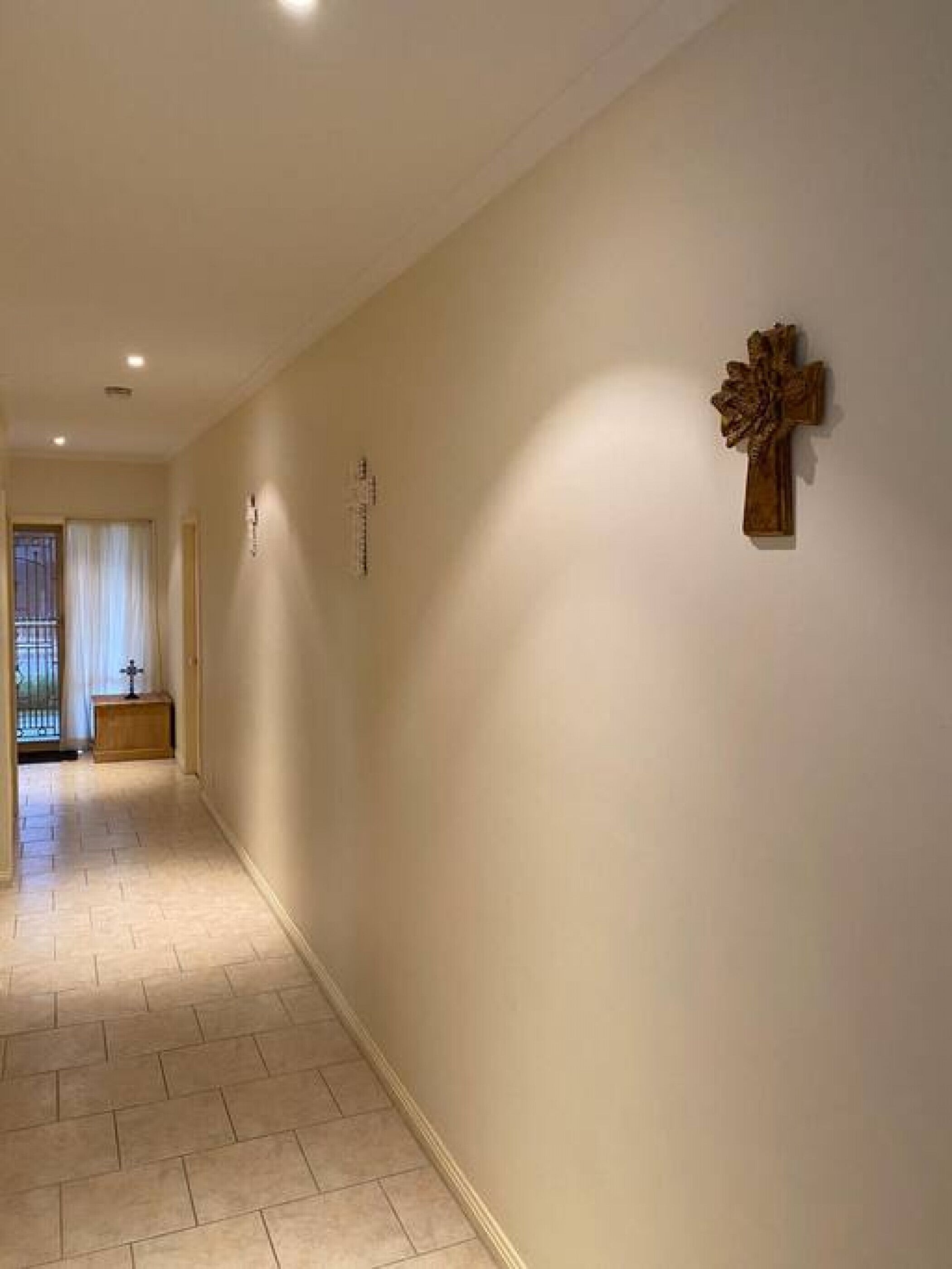 A hallway with a wooden cross on the wall, a small wooden cabinet, and a door leading to another room.