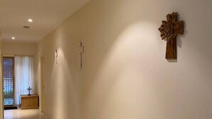 Un couloir orné d’une croix en bois au mur, un petit meuble en bois et une porte donnant sur une autre pièce.