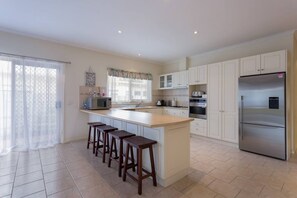 A kitchen with a large island, white cabinets, a stainless steel refrigerator, and a window with sheer curtains.