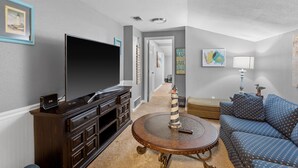 A living room with a flat-screen TV, dark wooden entertainment center, round coffee table, blue patterned sofa, and a hallway leading to another room. - Fun Summer Lake House w/Firepit located in Ft Worth, Beds 18+, next to Marina  (Fort Worth)