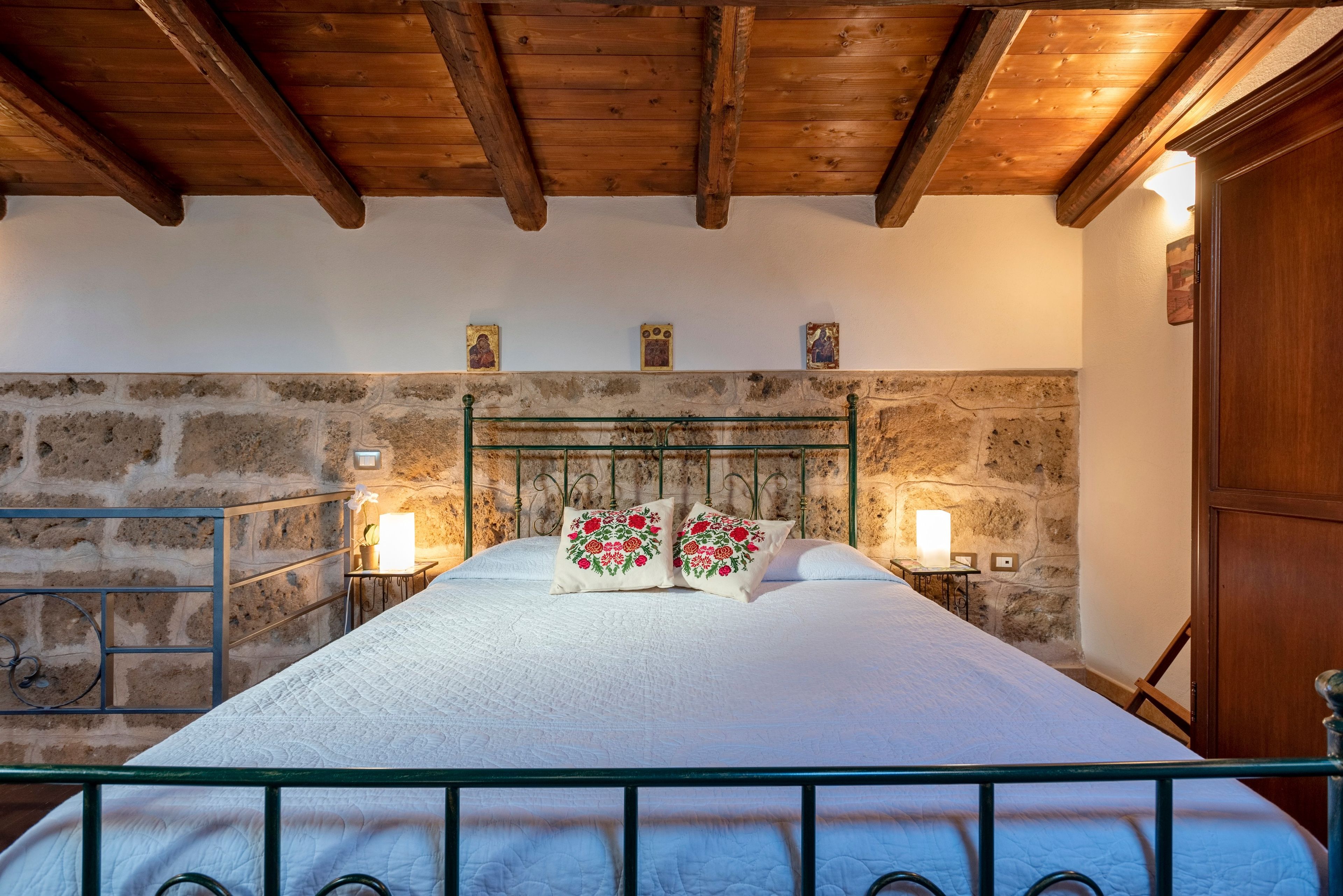 Ein Schlafzimmer mit Steinwand, Holzbalkendecke, einem Metallbettgestell und zwei Nachttischlampen.
