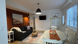 Una sala de estar moderna con comedor, que presenta un sofá negro, una mesa de comedor blanca y una alfombra estampada.