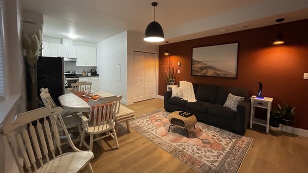 Una sala de estar con un sofá oscuro, una mesa de comedor con sillas, un tapete estampado y un cuadro en la pared.