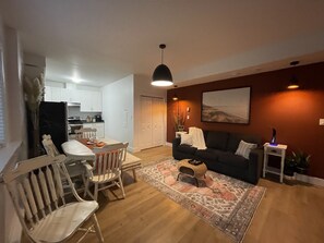 Una sala de estar con un sofá oscuro, una mesa de comedor con sillas, un tapete estampado y un cuadro en la pared.