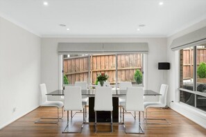 A modern dining area with a glass table and white chairs, a wooden fence view, and a large window. - Seagrove Escape - Near the beach & WIFI (Cowes)