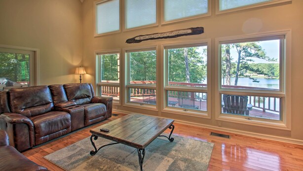 A spacious living room with a leather sofa, a wooden coffee table, and large windows offering a view of a lake and outdoor deck. - Private Dock & Pool Access! Hot Springs Home (Hot Springs)