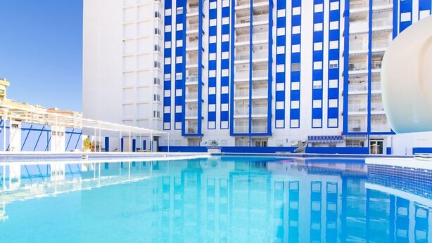 Una piscina con superficie de agua azul cristalina, rodeada por un edificio de rayas verticales azules y blancas.