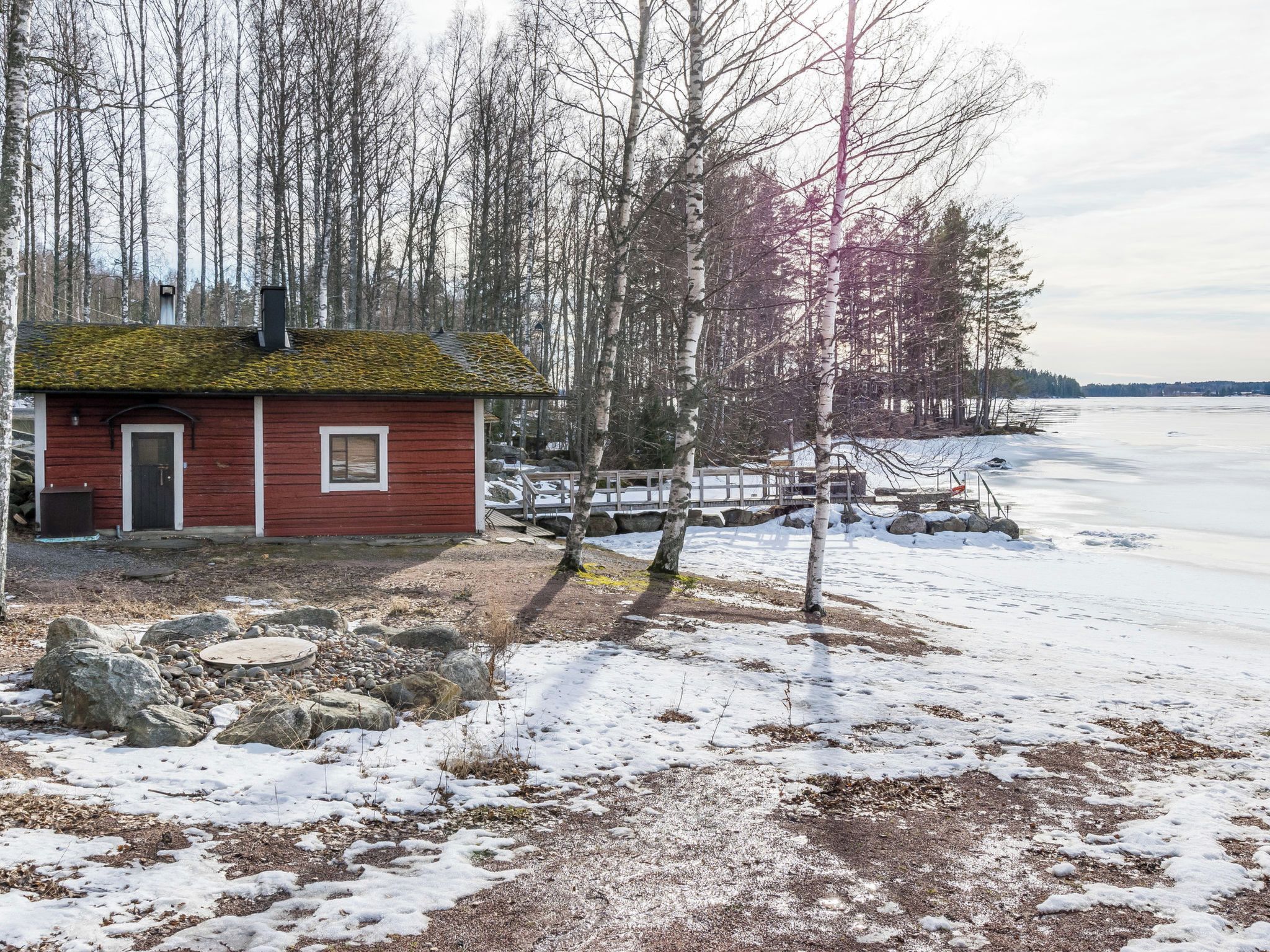 A small red cabin with a wooden deck and a dock extending into a frozen lake.