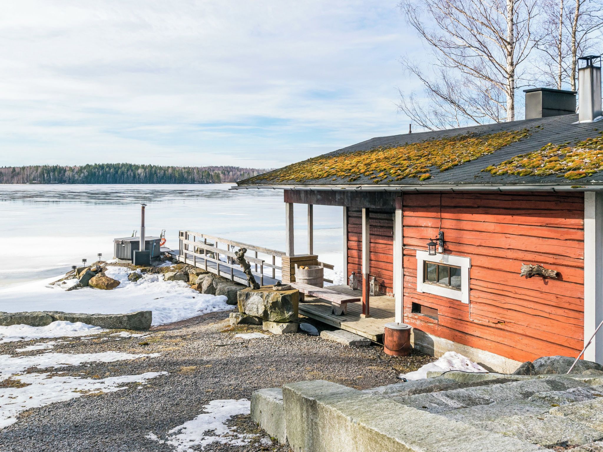 En röd trästuga med en brygga som sträcker sig ut i en frusen sjö, omgiven av stenar och snö.