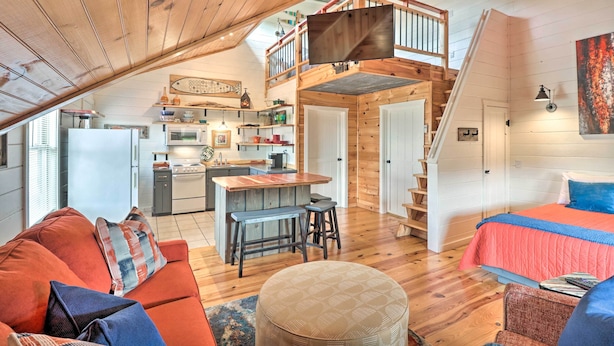 A cozy loft-style room with a wooden ceiling, a kitchen area, a dining space, and a bedroom with a bed and a window.
