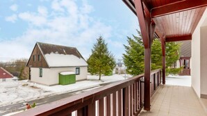 A balcony with a wooden railing overlooking a snowy landscape, a small house with a green door, and a clear sky.